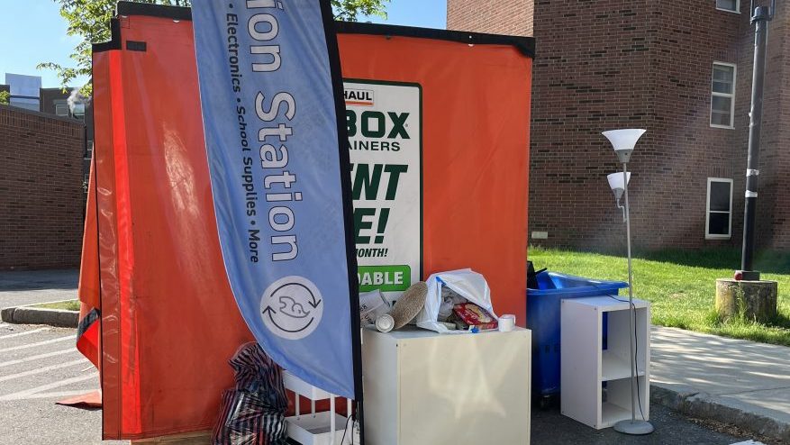 large donation box with assorted donated furniture items surrounding it outside in a parking lot. A large blue flag in front of the box reads "Donation Station"