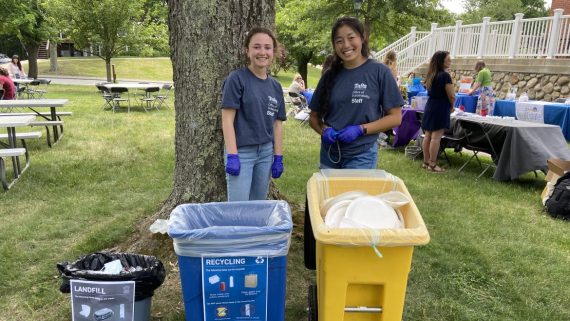 Two women stand facing the camera behind trash, recycling, and compost bins