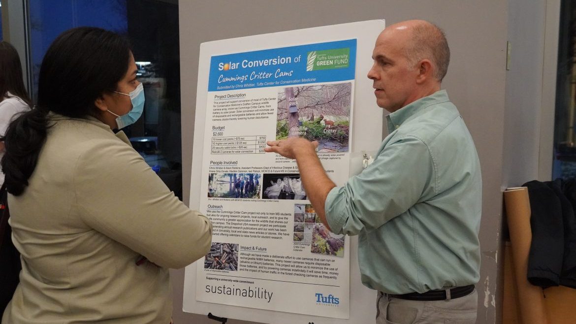 A man stands in front of a poster reading "Solar Conversion of Cummings Critter Cams" gesturing at the poster, explaining his Green Fund project to a woman 