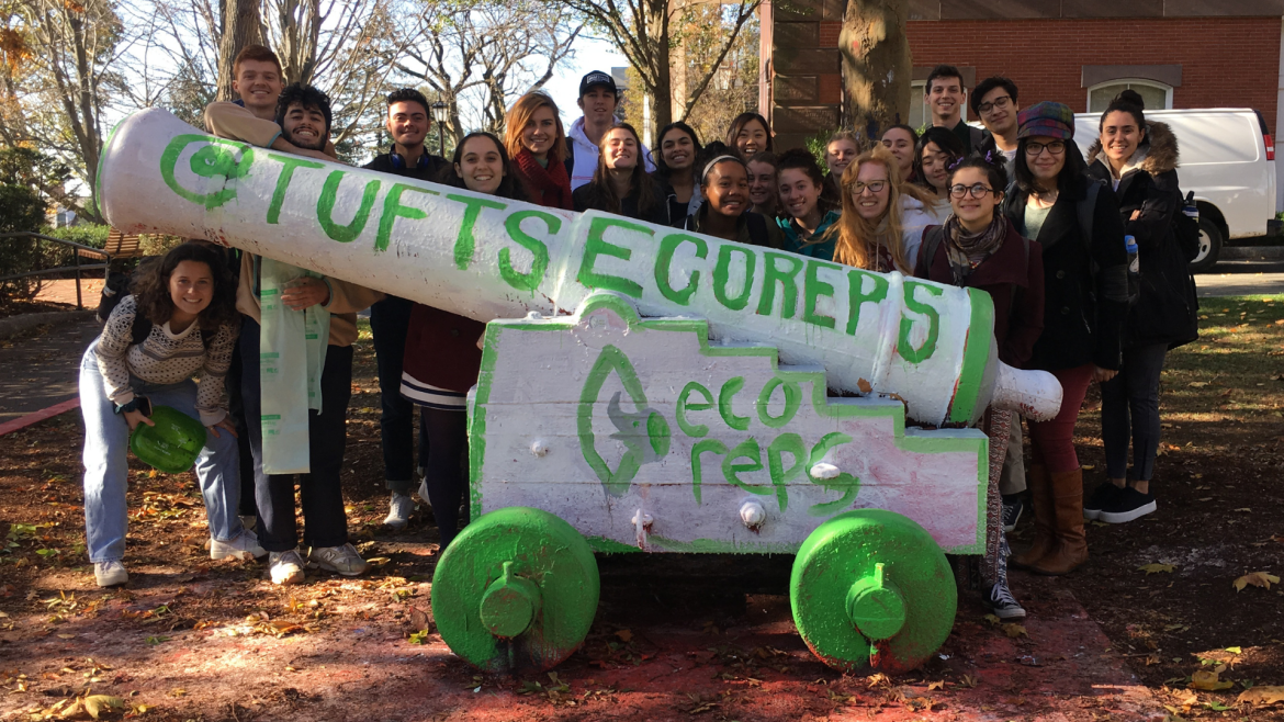 A group of about 20 students stands behind the Tufts cannon, which they have painted white and green. It reads "@Tufts Eco Reps" and has the Eco Reps logo painted.
