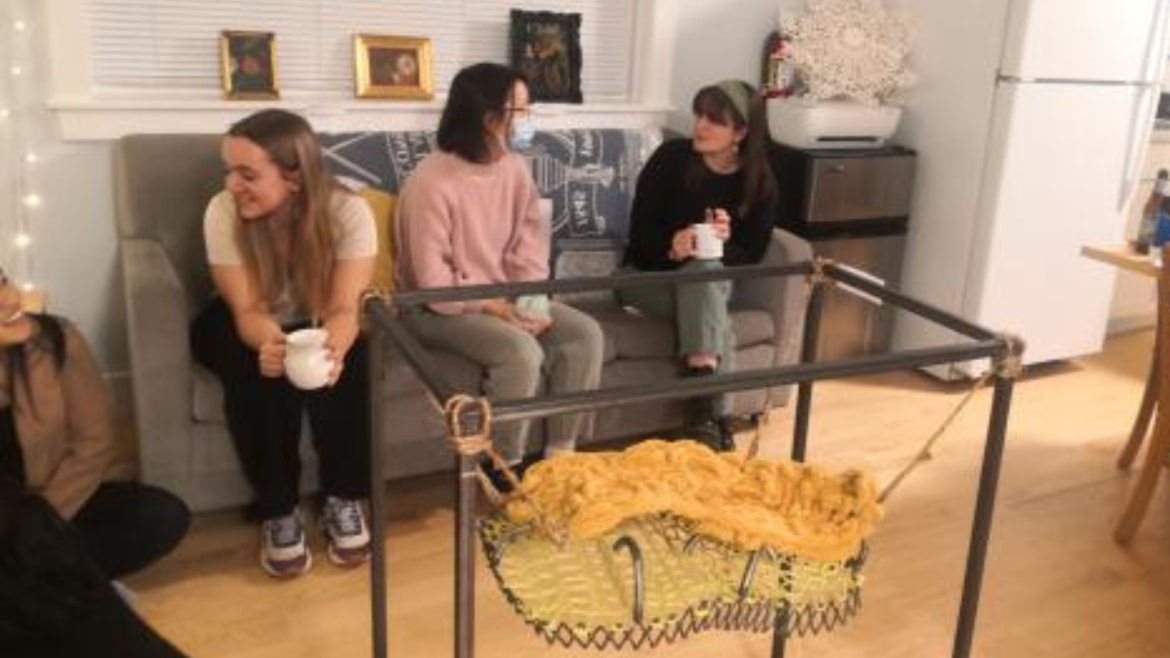Three students sitting on a couch talking with mugs in their hands. An art piece is front of them.