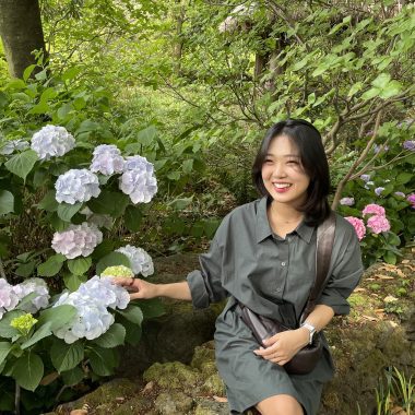 Woman sitting outside on a rock ledge next to blooming flowers and smiling at the camera.