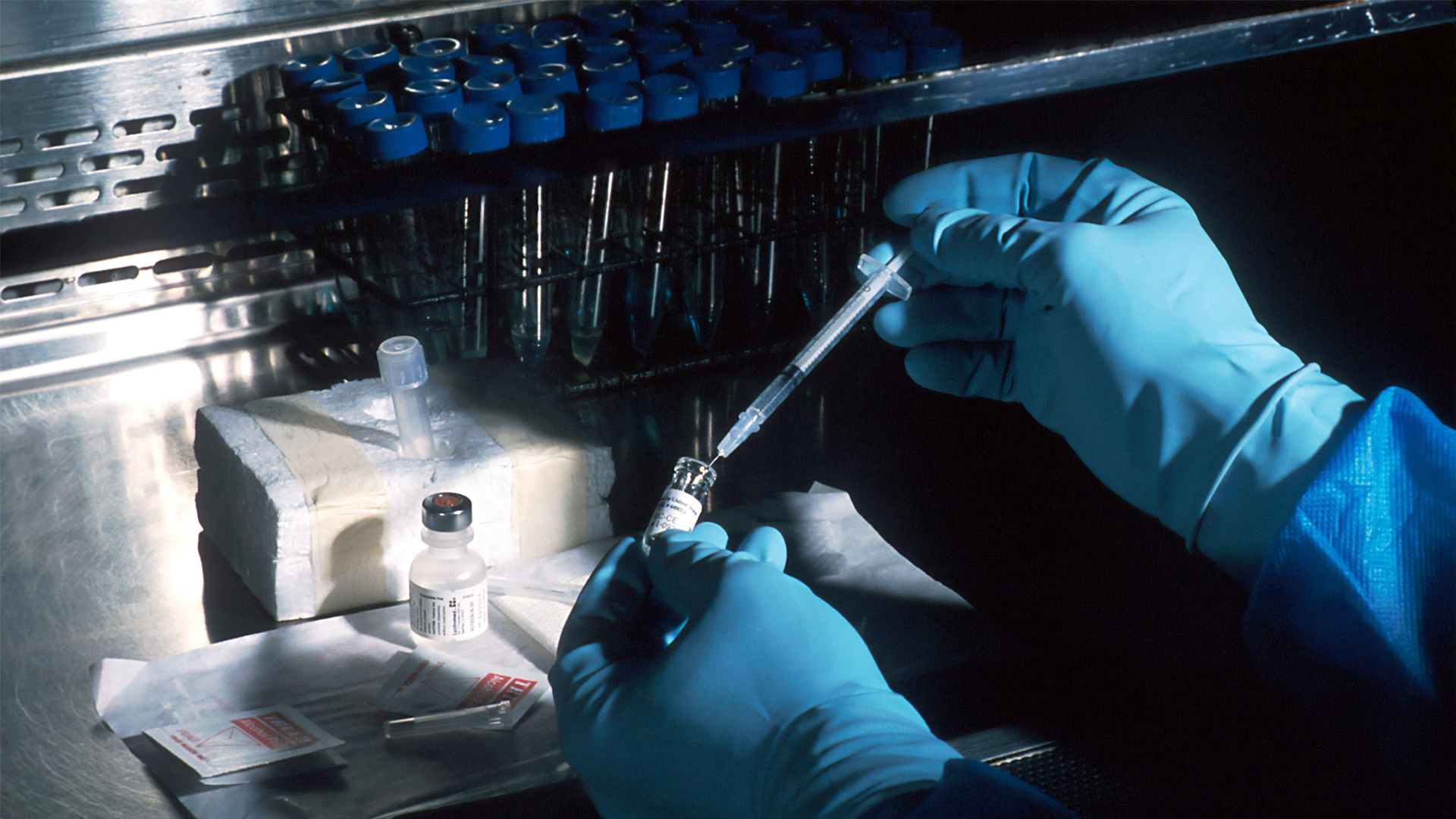 Gloved hands taking liquid out of a vial with a syringe. There are lab samples in the backgroun.