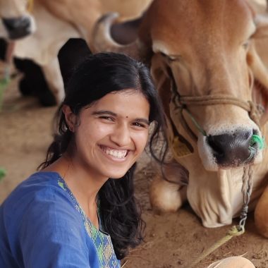 Prajna is wearing blue and smiling at the camera. She is seated on the ground. There is a cow seated next to her and behind her.