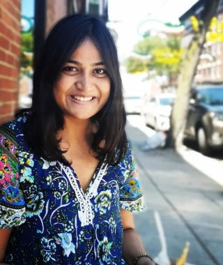 Half-body photo of Sulagna in a dark blue patterned shirt. She is sitting outside with the street sidewalk visible.